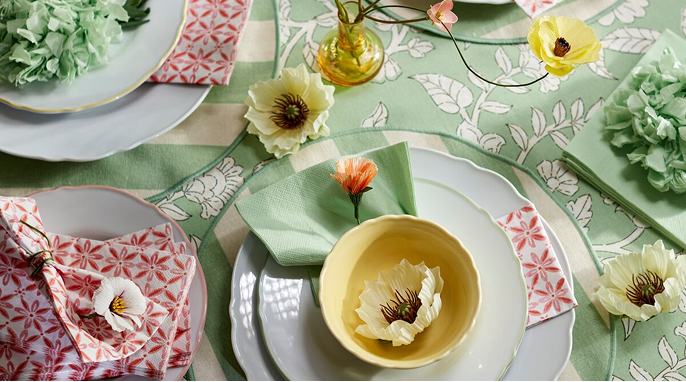 Svátečně prostřený jarní stůl se zeleným květovaným ubrusem SMÖRFISK a zeleno-bílým pruhovaným prostíráním SMÖRFISK, spolu se žlutou mísou UPPLAGA a bílými talíři a dezertními talířky UPPLAGA. Stůl je ozdobený umělými magnóliemi SMYCKA, papírovými ubrousky SMÖRFISK s červeno-bílým vzorem a zelenými ubrousky FANTASTISK.
