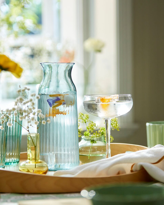 Světle tyrkysová karafa SMÖRFISK mezi dekorativními vázami a elegantním skleněným nádobím na stole.