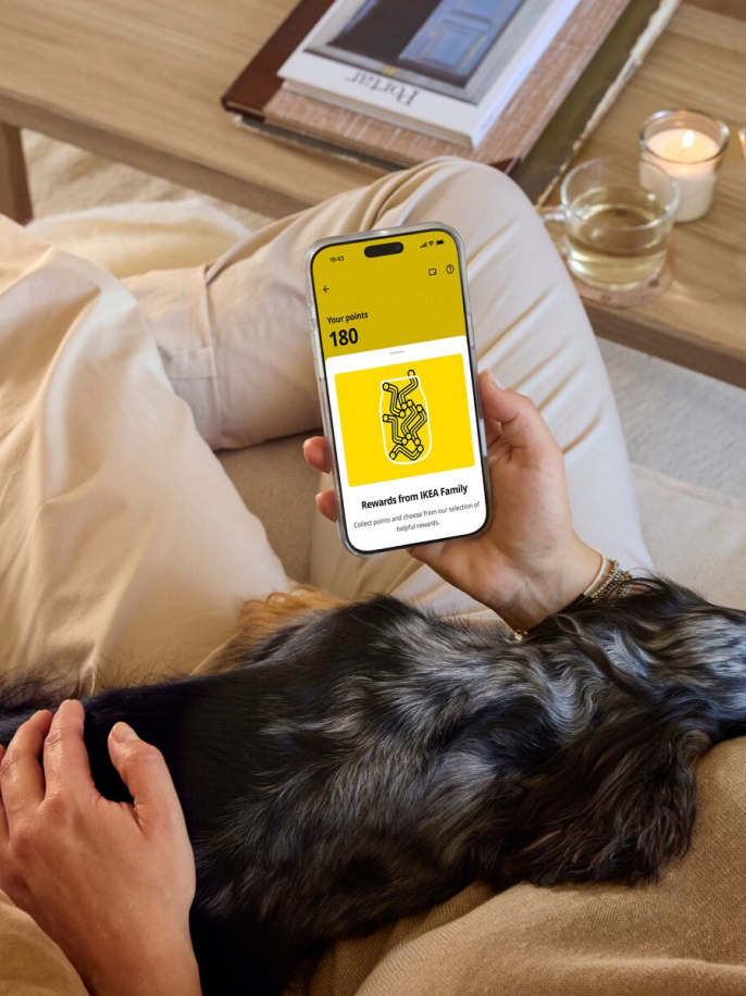Osoba leží na gauči, na klíně má psa a v ruce drží mobil s aplikací IKEA.