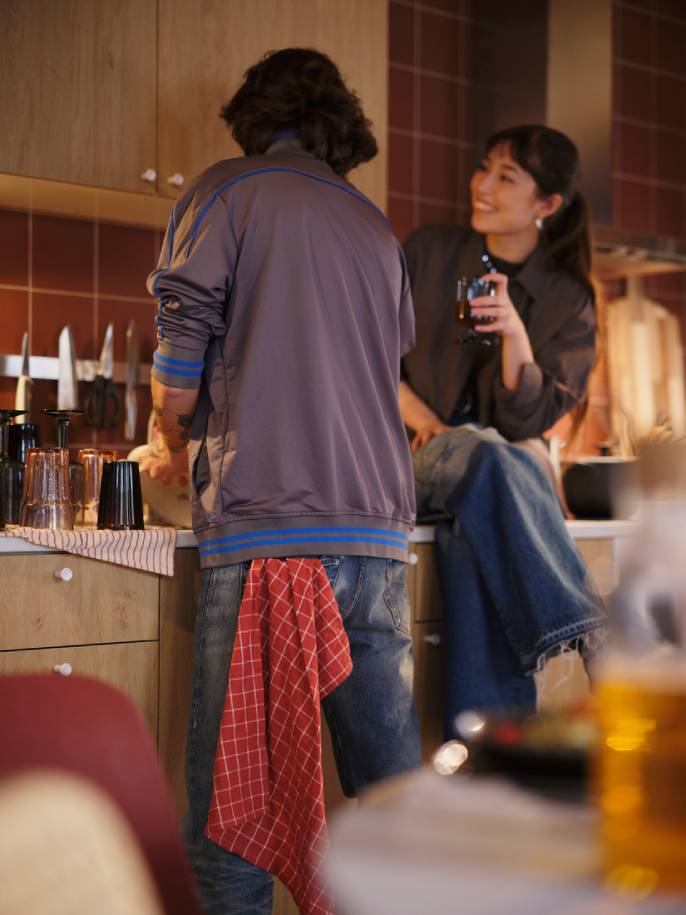 Mladý muž myje nádobí v dřezu, vedle něj sedí mladá žena se sklenkou.