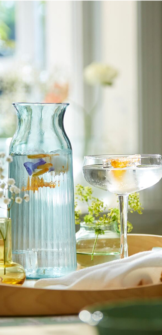 Světle tyrkysová karafa SMÖRFISK mezi dekorativními vázami a elegantním skleněným nádobím na stole.