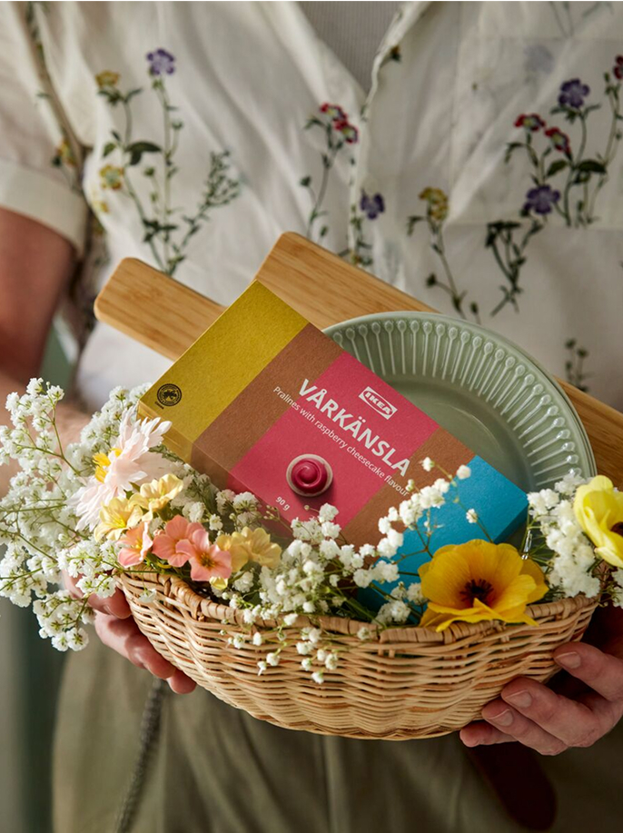 Osoba nese košík na pečivo SMÖRFISK jako dárkový koš, naplněný dárkovou krabičkou s pralinkami VÅRKÄNSLA a ozdobený umělými větvičkami vrby, zlatého deště a magnóliemi SMYCKA.