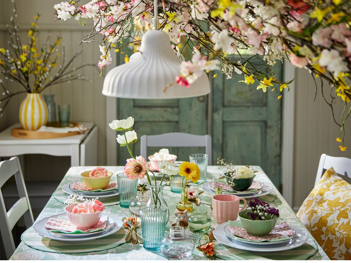 Slavnostně prostřený jarní stůl se žlutými, tmavě zelenými, růžovými a světle zelenými miskami UPPLAGA na dezertních talířcích a talířích UPPLAGA, zdobený umělými magnóliemi SMYCKA.